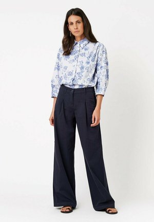 Woman wearing a blue and white floral button-up blouse with wide-leg navy trousers and black sandals, standing against a white background.