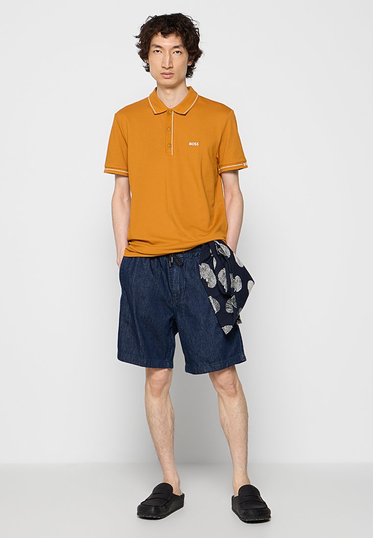 Polo shirt arancione con finiture bianche e logo, pantaloni corti di jeans e sciarpa a motivi. Le scarpe nere senza lacci completano l'outfit casual.