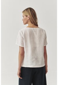 Hvid linned top med korte ærmer og en bred halsudskæring, parret med mørke bukser. Stoffet har en subtil tekstur.