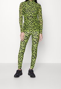 Czarny siatkowy body i legginsy z zielonym wzorem serc, z wysokim kołnierzem i dopasowanym kształtem, połączone z czarnymi butami sportowymi.