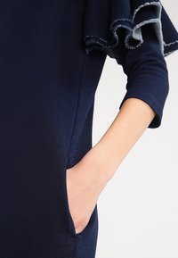 Granatowa sukienka z miękkiego materiału, z rękawami o długości trzy czwarte i falbaną na dole. Widoczny detal kieszeni po lewej stronie.