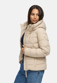Beige pufferjacka med hög krage och dragkedjestängning. Har ett horisontellt quiltat mönster och sidofickor. Bärs över en marinblå topp och jeans.