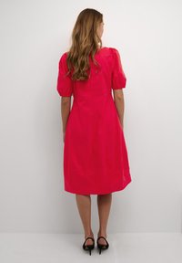 Ružičasta haljina do koljena s nabranim rukavima i strukom koji ističe figuru. Izrađena od glatkog materijala s klasičnim siluetom. Nošena s crnim salonkama.