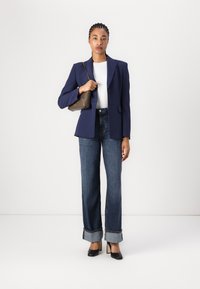 Marineblazer über einem weißen Oberteil, kombiniert mit hoch taillierten, weiten Jeans und spitzen schwarzen Absätzen. Trägt eine braune Handtasche.
