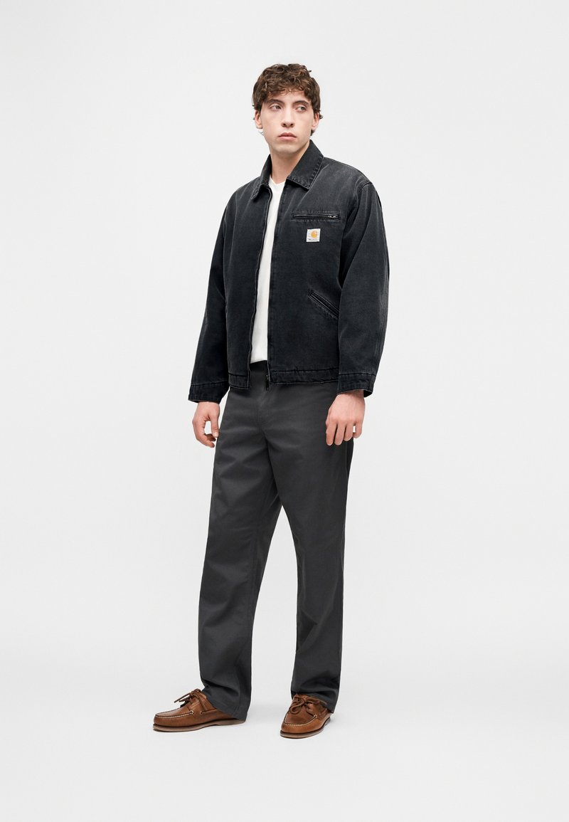 Giacca in denim nera con cerniera frontale, tasca applicata, abbinata a pantaloni grigi e scarpe marroni. Design semplice a taglio dritto.