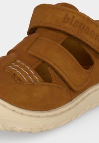 Sandale bébé en daim marron avec deux brides Velcro et détails de couture blanche sur une semelle en caoutchouc blanche.