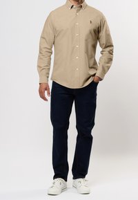 Beiges Hemd mit Knöpfen, langen Ärmeln und Kragen, kombiniert mit dunkelblauen Jeans und weißen Turnschuhen. Verfügt über eine Logosstickerei auf der Brust.