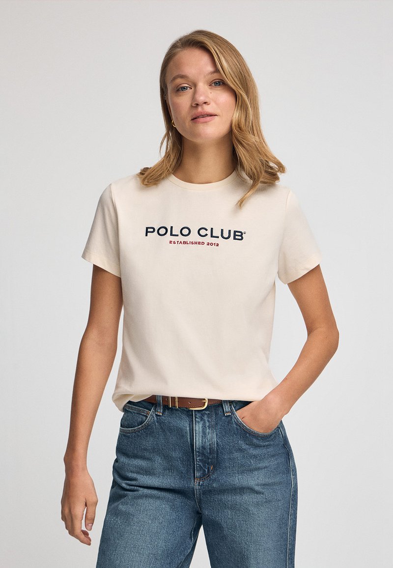 Beige bomulls-T-shirt med korta ärmar, rund halsringning, med texten "POLO CLUB" i marinblått och rött, kombinerad med blå denimjeans.