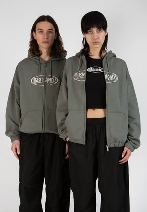 Green zip-up hoodies with a logo on the front, paired with black loose-fitting pants. Hoodies have elastic cuffs and front pockets.