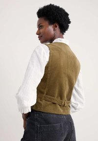 Olivenozelena kordura prsluk prilega obliku, s dugim gumbima na leđima i teksturiranom površinom, složen preko bijele košulje s dugim rukavima.