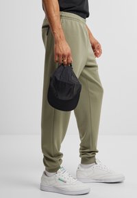Pantalons de jogger vert olive clair avec une taille à cordon et une poche latérale, associés à des baskets blanches. Une casquette noire est tenue à la main.
