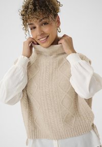 Knit beige sweater with a high ribbed collar, textured cable pattern, and loose fit. Underneath, a white billowed sleeve shirt is visible.