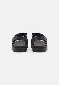 Zwarte sandalen met brede banden van synthetisch materiaal, voorzien van gouden accenten en een platte, gestructureerde zool. Ontworpen voor comfort en ondersteuning.