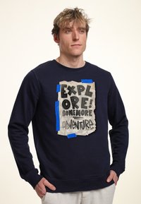 Felpa navy con collo a giro, caratterizzata da una stampa grafica su carta testurizzata che ricorda un materiale strappato, con nastro blu come accentuazione.
