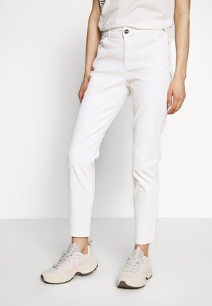 Person iført tætsiddende hvide jeans og beige atletik sneakers, stående mod en ensfarvet hvid baggrund.