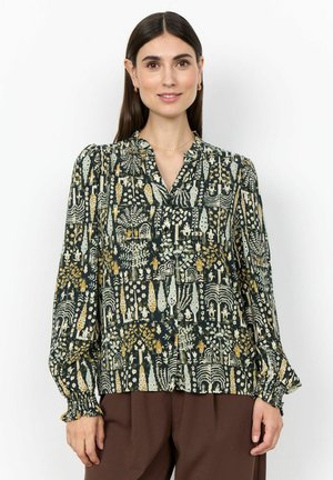 Frau trägt eine langärmelige, knöpfbare Bluse mit einem mehrfarbigen abstrakten Blumenmuster und braune Hose, steht vor einem schlichten Hintergrund.