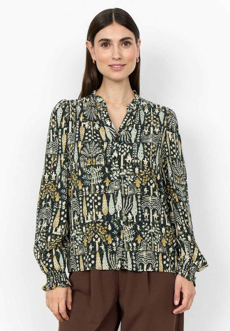 Femme portant un chemisier boutonné à manches longues avec un motif floral abstrait multicolore et un pantalon marron, debout devant un fond uni.