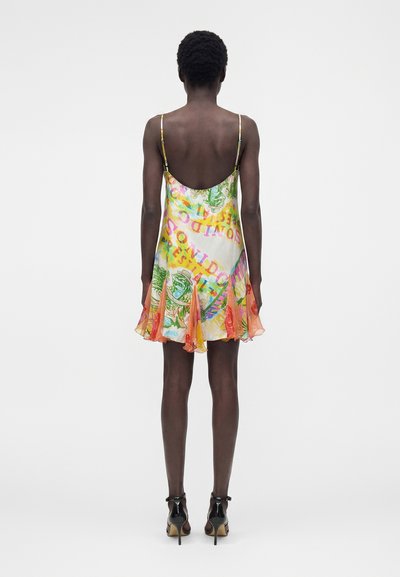 Άτομο με κοντά σγουρά μαλλιά που φοράει πολύχρωμο αμάνικο μίνι φόρεμα με αφηρημένα σχέδια και μαύρα σανδάλια με ψηλό τακούνι, στραμμένο προς τα πίσω.