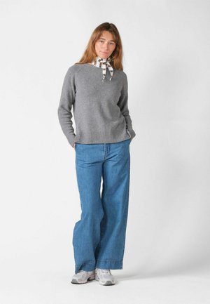 Kvinde iført grå sweater, blå vidde jeans, hvide sneakers og ternet halstørklæde, stående med hænderne i lommerne mod en hvid baggrund.