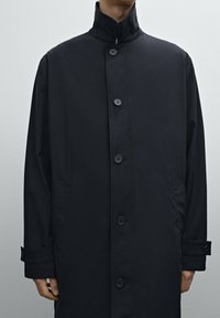 Schwarzer, langärmeliger Mantel mit hohem Kragen, Knopfschluss und Seitentaschen. Hergestellt aus glattem, leichtem Material mit matter Oberfläche.