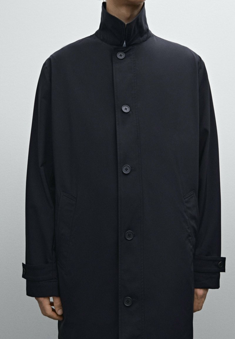 Schwarzer, langärmeliger Mantel mit hohem Kragen, Knopfschluss und Seitentaschen. Hergestellt aus glattem, leichtem Material mit matter Oberfläche.