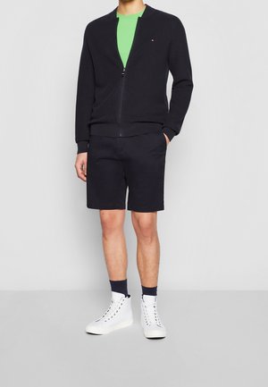 Granatowy sweter zapinany na zamek z teksturowanej tkaniny, zestawiony z czarnymi szortami i białymi butami sportowymi typu high-top. Zielona koszulka pod spodem, granatowe skarpetki.