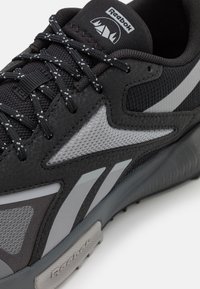 Sneakers neri con tomaia in pelle sintetica, accenti in rete, strisce riflettenti grigie e lacci bianchi. Presentano una suola in gomma testurizzata e un logo.