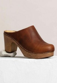 Bruine leren klomp met een ronde neus, houten hak en zichtbare stiksels langs de rand. Geplaatst op een gestructureerd witte ondergrond.