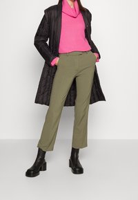 Cappotto nero trapuntato sopra un maglione rosa a coste e pantaloni verdi oliva su misura, abbinati a stivaletti neri con tacco spesso.