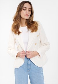 Krémový dvouřadý blazer se zlatými knoflíky, texturovaná tkanina a klopové límce, nošený přes bílý top a vysoké modré džíny.