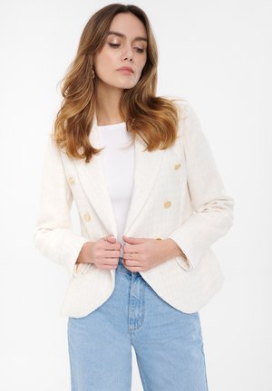 Krémový dvouřadý blazer se zlatými knoflíky, texturovaná tkanina a klopové límce, nošený přes bílý top a vysoké modré džíny.