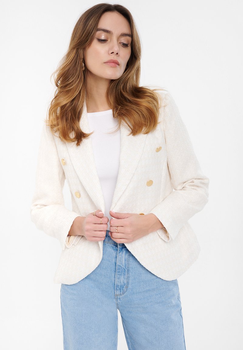 Krémový dvouřadý blazer se zlatými knoflíky, texturovaná tkanina a klopové límce, nošený přes bílý top a vysoké modré džíny.