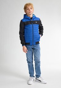 Giacca trapuntata blu con maniche e cappuccio neri, con zip frontale, polsini a costine, indossata sopra una camicia azzurro chiaro e abbinata a jeans di denim e sneaker.