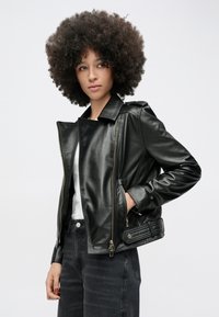 Göndör hajú nő fekete bőrdzsekit visel fehér felső és sötét farmer fölött, az egyik kezét a dzseki zsebében tartva pózol.