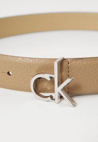 Cintura in pelle marrone chiaro con finitura testurizzata, fibbia in metallo argento con logo "ck", con cuciture visibili e fori per la regolazione.