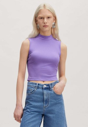 Junge Frau mit langen blonden Haaren, die große silberne Ohrringe trägt, ein ärmelloses geripptes lilafarbenes Crop-Top und hoch taillierte blaue Jeans mit einer Hand in der Tasche.