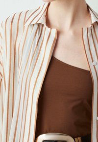 Lightweight, sheer white shirt with vertical brown stripes, buttoned collar, layered over a brown tank top. Features a thick belt buckle.