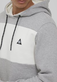 Grijze en witte hoodie met een driehoekig logo, trekkoord met zilveren accenten en textuurstof. Kleurblok ontwerp.