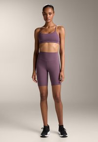 Hajcsíkos mályva sportmelltartó vékony pántokkal és magas derekú sorttal. Simaságú, rugalmas anyag. A modellen fekete sportcipők vannak. Semleges háttér.
