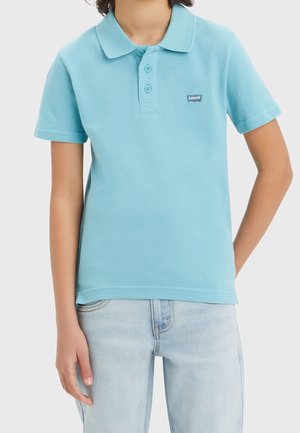 Enfant portant un polo bleu clair à manches courtes avec trois boutons et un jean clair, debout avec une main derrière le dos.