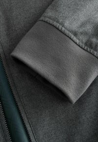 Graue Zip-jacke aus strukturiertem Stoff. Verfügt über einen gerippten Bund und ein glattes Innenfutter mit geometrischem Muster.