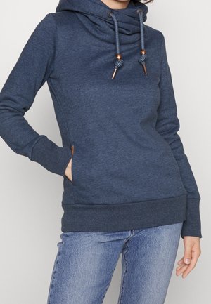 Frau trägt einen blauen Kapuzenpullover mit Kordelzügen und Vordertaschen, kombiniert mit hellblauen Jeans, steht mit einer Hand in der Tasche.