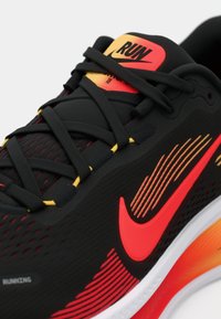 Μαύρο αθλητικό παπούτσι με υφασμάτινη επιφάνεια, κόκκινες και κίτρινες λεπτομέρειες, έντονο λογότυπο swoosh και σόλα με γραadiεντόνιο που περνά από το λευκό στο πορτοκαλί.