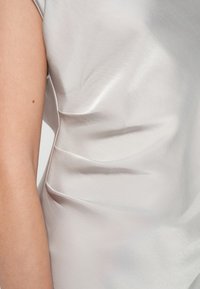 Blouse en soie de couleur argent pâle, présentant de doux plis sur le côté et une texture lisse et brillante, mettant en valeur un design fluide.