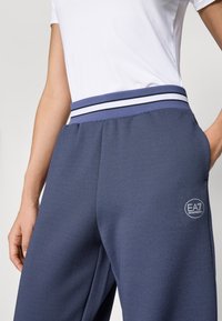 Marine idrettsbedrakter med teksturert stoff, med et flerfarget stripete liv og en brodert EA7-logo på benet.