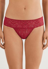 Tanga en dentelle rouge profond avec un motif floral. Présente une taille en dentelle festonnée et un tissu lisse pour le confort.