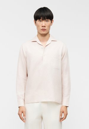 Lichtroze shirt met lange mouwen van een gestructureerde stof, met een geknoopte kraag en een borstzak, gedragen over beige broek.