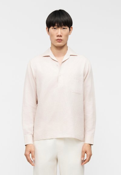 Chemise rose clair à manches longues en tissu texturé, avec un col boutonné et une poche poitrine, portée avec un pantalon beige.