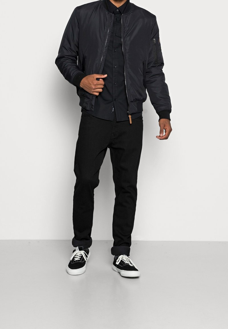 Giacca bomber nera con polsini a coste, tasche con zip e finitura liscia, abbinata a camicia nera con bottoni e jeans scuri. Sneakers nere.