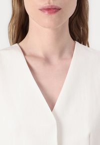 Bijela majica s V izrezom izrađena od teksturiranog materijala, s čistim rezom i suptilnim, mekim sjajem. Izrez je istaknut, stvarajući minimalistički izgled.